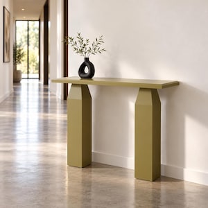 May include: A mustard yellow console table with a rectangular top and two thick, tapered pillar-style legs. A black, hollow, rounded vase containing green leafy branches sits centered on the flat surface of the table.