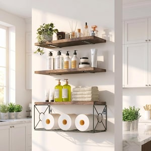May include: Three wooden shelves with dark metal brackets are mounted on a white wall. The shelves hold various bathroom items, including bottles, jars, towels, and toilet paper rolls. A small potted plant sits on the top shelf.