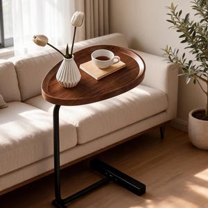 May include: A dark brown, oval-shaped side table with a black metal base. The table holds a white teacup on a wooden coaster, a white vase with three white tulips. The table is next to a beige sofa.