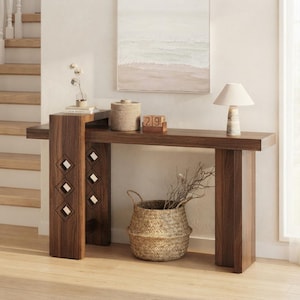 May include: A dark wood console table with a tiered design and diamond-shaped cutouts on the left support. The table holds a small lamp, a lidded container, and a wooden block calendar showing the number 29. A woven basket sits underneath.