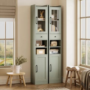 May include: Two tall, sage green storage cabinets featuring a vertical design with a glass-paneled upper door, a single drawer, an open middle shelf, and a solid lower door. Each cabinet is equipped with black metal handles and knobs.