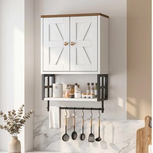 May include: A white kitchen cabinet with two doors and a brown top, mounted above a shelf with a black metal frame. Below the shelf is a hanging rack with kitchen utensils and a towel. The cabinet doors have a decorative design.