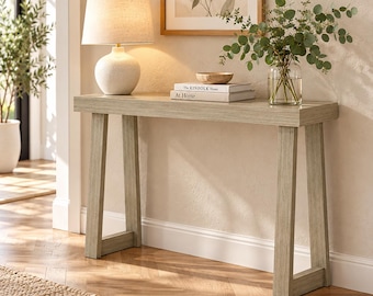May include: A light-toned wooden console table with A-frame legs. On the surface sits a white textured table lamp, a small wooden bowl, and two stacked books titled The Kinfolk Home and At Home. A clear glass vase with green eucalyptus branches.