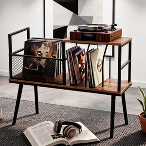 May include: A wooden record player stand with black metal frame. The stand has two shelves, one with a record player and the other with vinyl records. A wire mesh basket holds more records. Headphones and a book are on the floor.