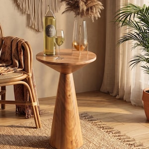 May include: A round wooden side table with a conical base. On the table sits a tall clear glass bottle filled with a golden liquid, a clear stemmed wine glass containing a golden liquid, and a clear glass vase holding dried pampas grass stems.