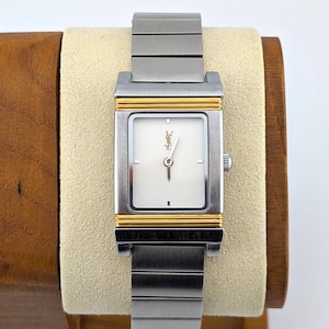 May include: A rectangular wristwatch with a silver-tone metal link band and a white face. The watch features gold-tone accents on the top and bottom of the case, with the Yves Saint Laurent logo positioned at the twelve o'clock marker on the dial.