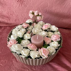 Peut inclure: Composition florale avec une bougie rose entourée de roses blanches et roses. Les fleurs sont disposées dans un récipient décoratif marron clair à bords festonnés, avec des accents de mousse verte.