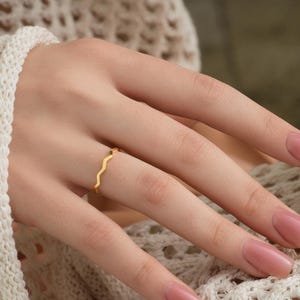 May include: A gold-colored ring with a wavy design is worn on a finger. The ring is thin and delicate. The hand is resting on a cream-colored knitted sweater. The nails are painted with a light pink nail polish.