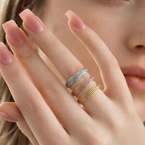 May include: Three rings with a leaf design are displayed on a finger. The rings are silver, rose gold, and gold. The rings are stacked on the finger. The background is blurred.