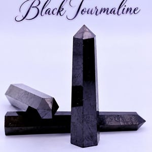 May include: Three black tourmaline crystals shaped into pointed towers. One crystal stands vertically in the foreground, while two others lie horizontally behind it. The text Black Tourmaline is written in a dark script font above the crystals.