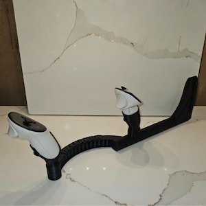 May include: A black and white VR controller stand. The stand has a curved, textured arm with two white controllers attached. The controllers have black buttons and are angled towards the viewer. The stand is on a white surface.