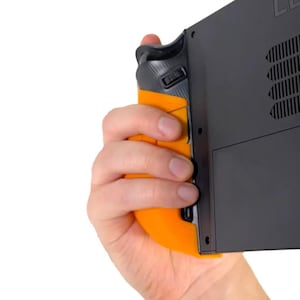 May include: An orange and black gaming controller grip is attached to a black handheld gaming device. The grip has an ergonomic design with a textured surface for enhanced control. The device is held against a white background.