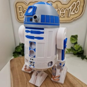 Può includere: Una statuetta di R2-D2 stampata in 3D, con corpo bianco e dettagli blu e argento. Il droide di Star Wars è posizionato su una superficie di legno. La statuetta presenta dettagli ed è un oggetto da collezione.