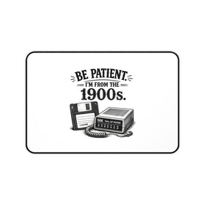 Puede incluir: Una alfombrilla de escritorio rectangular con un gráfico en blanco y negro de un disquete de 3,5 pulgadas y un módem de acceso telefónico de 56K con cable en espiral. El texto dice: BE PATIENT. I'M FROM THE 1900s. Diseño centrado en fondo blanco.