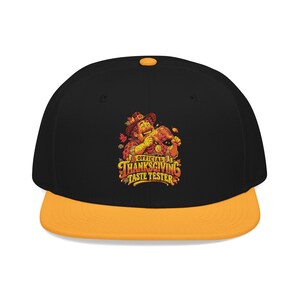 May include: A black baseball cap with an orange brim and top button. The front features a graphic of a person in a hat eating a turkey leg, surrounded by autumn leaves, with the text "OFFICIAL THANKSGIVING TASTE TESTER" in orange and yellow.