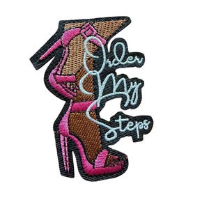 Peut inclure: Un écusson brodé représentant une chaussure à talons hauts dans des tons rose et marron, avec le texte bleu clair "Order my steps" cousu par-dessus. L'écusson présente une bordure noire et un travail de fil détaillé sur l'ensemble.