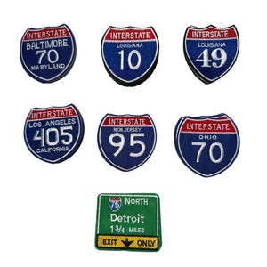 Puede incluir: Un conjunto de parches bordados con señales de autopistas interestatales. Los parches muestran los números de ruta y los nombres de los estados, incluyendo Baltimore, Maryland; Luisiana; Los Ángeles, California; Nueva Jersey y Ohio. Un letrero verde indica una salida en Detroit.