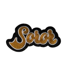 May include: Embroidered patch with the word "Saint" in gold script lettering. The text is outlined in white and set against a black background. The patch has a decorative, wavy border.