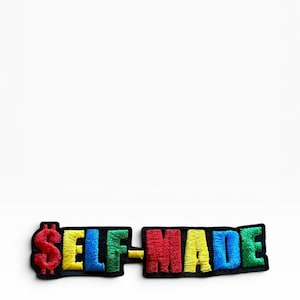 May include: An embroidered patch featuring the text "SELF-MADE" with a dollar sign replacing the letter S. The letters are stitched in vibrant red, yellow, blue, and green thread, each outlined in black against a solid black background.