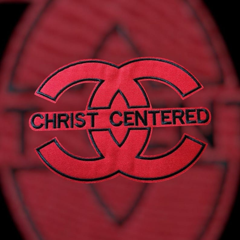 Puede incluir: Un parche bordado de color rojo con un dise&ntilde;o estilizado de dos letras C entrelazadas con un borde negro. El texto "CHRIST CENTERED" est&aacute; bordado en letras may&uacute;sculas negras en el centro del dise&ntilde;o, dividiendo horizontalmente las dos formas.