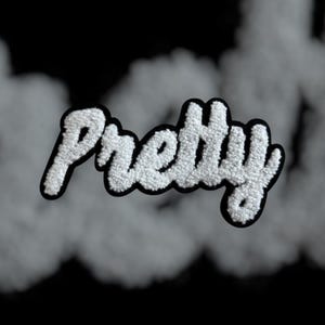 May include: The word "Pretty" in a white, textured font with a black outline, set against a smoky gray background. The letters appear to have a fuzzy, plush texture, creating a 3D effect. The background is a gradient of gray and black.