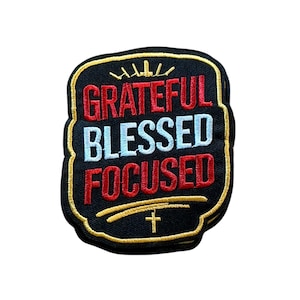 May include: A black embroidered patch with a gold border featuring the text GRATEFUL in red, BLESSED in light blue, and FOCUSED in red. Above the text is a gold crown icon, and below the text is a gold curved line with a small cross symbol.