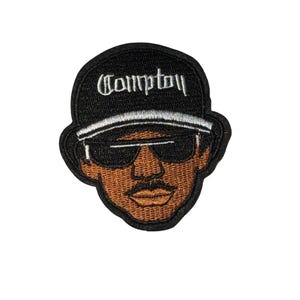 May include: Embroidered patch featuring a stylized face wearing sunglasses and a cap. The cap is black with the word "Compton" in white script. The face is brown with a black mustache and sunglasses.