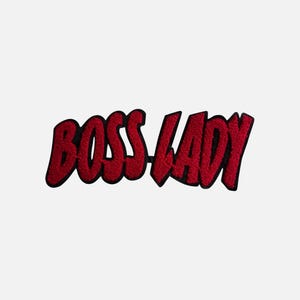 Può includere: Una toppa ricamata rossa con le parole "BOSS LADY" in un carattere audace in stile fumetto. Il testo ha un contorno nero e la toppa è su uno sfondo bianco.