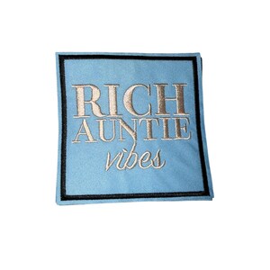 May include: A light blue square patch with the embroidered text "RICH AUNTIE vibes" in gold thread. The patch is outlined with a black border.