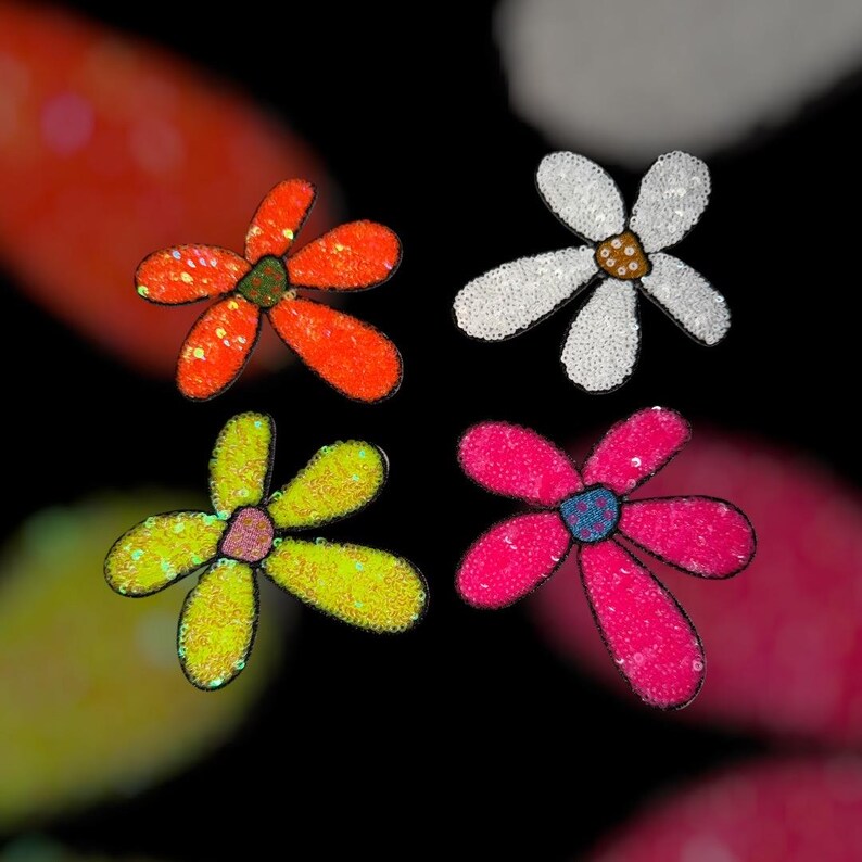 Puede incluir: Cuatro apliques de flores con lentejuelas, naranja, blanco, amarillo y rosa. Cada flor tiene un centro de diferente color. Los p&eacute;talos est&aacute;n delineados en negro, sobre un fondo negro con colores borrosos.
