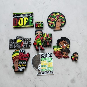 May include: A collection of colorful shoe charms with Black history and culture themes. The charms feature phrases like "Unapologetically Dope," "I Am Black History," and images of Black women and the Juneteenth holiday.