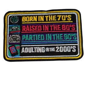 May include: Embroidered patch with a black background and a yellow border. The patch features text in various colors: "BORN IN THE 70'S", "RAISED IN THE 80'S", "PARTIED IN THE 90'S", and "ADULTING IN THE 2000'S".