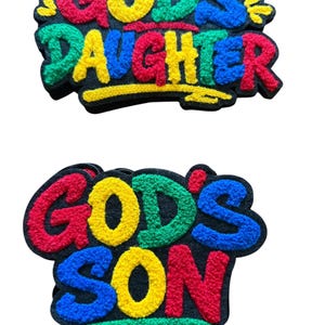Può includere: Due toppe in tessuto trapuntato con supporto nero. La toppa superiore presenta il testo "GOD'S DAUGHTER" in lettere testurizzate multicolori con una piccola corona gialla. La toppa inferiore presenta il testo "GOD'S SON" in lettere testurizzate simili.