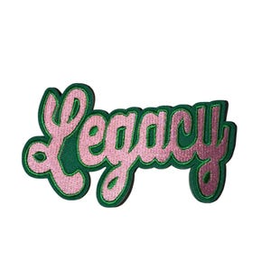 May include: A green and pink embroidered patch with the word "Legacy" in a cursive font. The word is in pink with a green outline and a green background. The patch is on a white background.