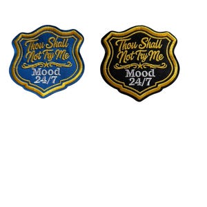 Peut inclure: Deux écussons brodés avec un motif de bouclier. L'un est bleu avec une bordure dorée et le texte "Thou Shall Not Try Me" et "Mood 24/7". L'autre est noir avec une bordure dorée et le même texte.