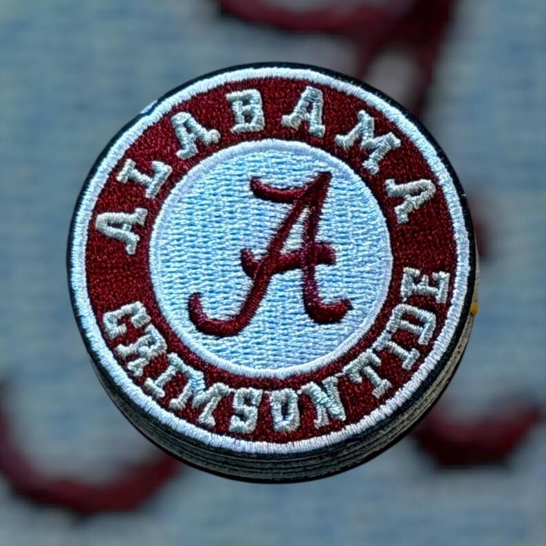 Peut inclure: Un &eacute;cusson brod&eacute; circulaire avec une bordure rouge cramoisi portant le texte blanc ALABAMA CRIMSON TIDE. Le centre est un cercle bleu clair avec une lettre A en &eacute;criture cursive rouge cramoisi fonc&eacute;. L'&eacute;cusson a une bordure ext&eacute;rieure noire.