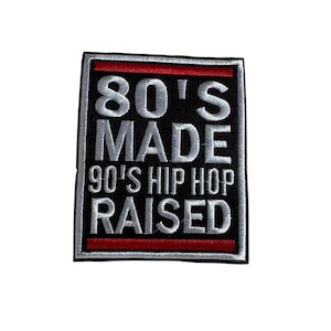 May include: A square patch with the text "80'S MADE 90'S HIP HOP RAISED" in silver embroidery on a black background. The patch has a red stripe at the top and bottom, and a silver border.