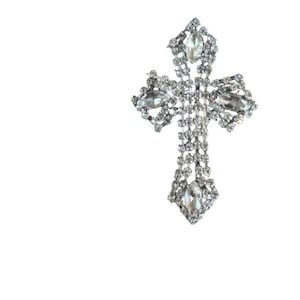 May include: A silver-toned cross brooch featuring a grid of small clear rhinestones and four larger marquise-cut clear crystals positioned at the top, bottom, and both sides of the cross structure, creating a reflective and ornate appearance.