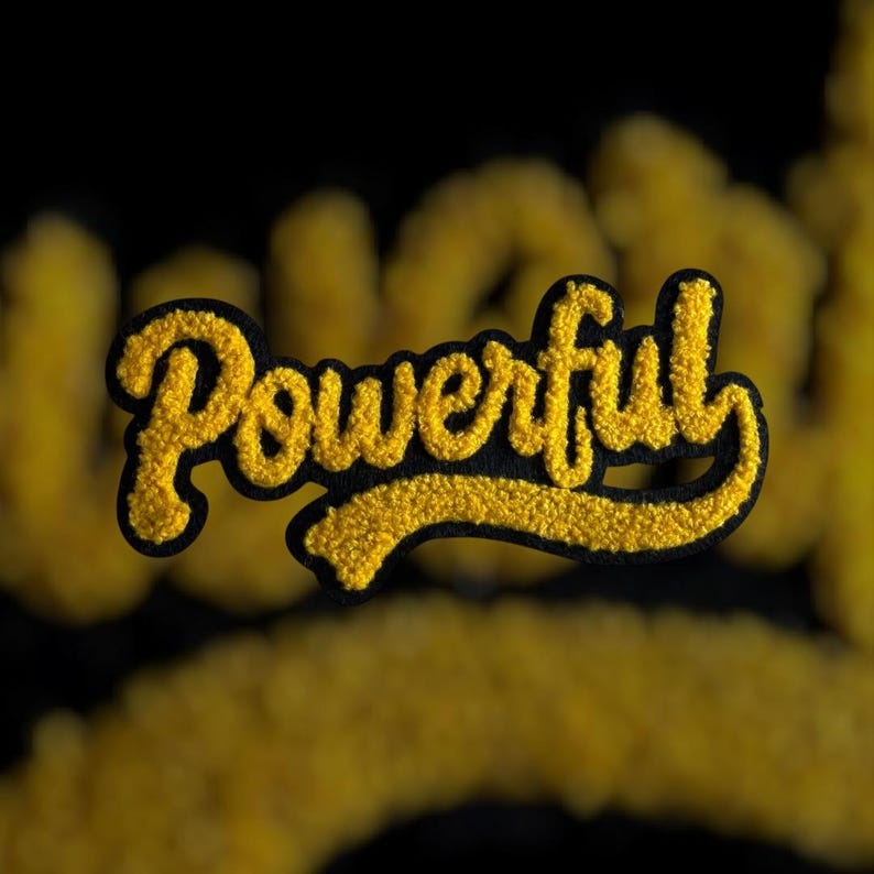 K&ouml;nnte beinhalten: Ein gelber Chenille-Aufn&auml;her mit dem Wort "Powerful" in einer Schreibschrift, schwarz umrandet. Der Aufn&auml;her hat ein strukturiertes, flauschiges Aussehen vor einem verschwommenen schwarzen Hintergrund.