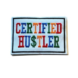 Puede incluir: Un parche rectangular bordado con fondo blanco y borde negro. El texto "CERTIFIED HUSTLER" está cosido en letras de bloque multicolores, con un signo de dólar verde que reemplaza la letra S en la palabra HUSTLER.