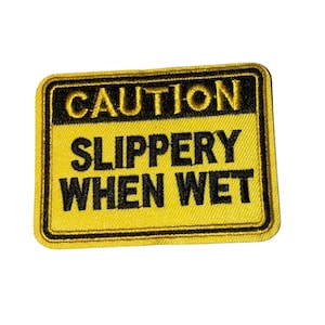 May include: A yellow and black embroidered patch with the words "CAUTION SLIPPERY WHEN WET". The patch has a black border and is rectangular in shape.