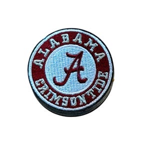 May include: A circular embroidered patch featuring a light blue center with a dark red script letter A. The border is dark red with the white text ALABAMA CRIMSON TIDE encircling the center. The entire patch has a white stitched edge.