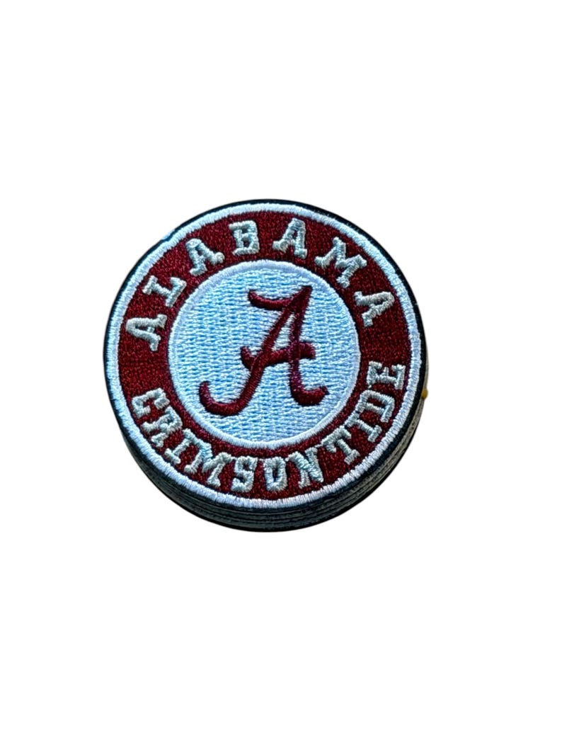 Peut inclure: Un &eacute;cusson brod&eacute; circulaire pr&eacute;sentant un centre bleu clair avec une lettre A en &eacute;criture rouge fonc&eacute;. La bordure est rouge fonc&eacute; avec le texte blanc ALABAMA CRIMSON TIDE entourant le centre. L'&eacute;cusson a un bord cousu blanc.
