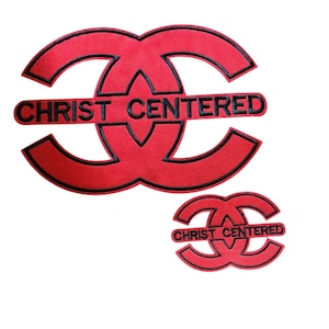 Peut inclure: Deux écussons brodés rouges avec des bordures noires, chacun présentant un motif stylisé de deux C entrelacés. Le texte CHRIST CENTERED est brodé en noir au centre de chaque écusson, positionné horizontalement sur les formes entrelacées.
