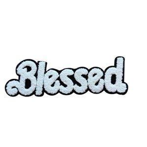 Könnte beinhalten: Ein weißer, flauschiger Aufnäher mit dem Wort "Blessed" in einer kursiven Schriftart. Die Buchstaben sind mit einem weichen, strukturierten Material gefüllt und schwarz umrandet. Der Aufnäher ist zum Aufnähen oder Aufbügeln auf Stoff gedacht.