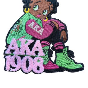 May include: An embroidered patch featuring a cartoon character with black hair, green eyes, and gold hoop earrings. The character wears a green and pink jacket with the text AKA, black trousers, and green boots. The text AKA 1908 appears in pink.