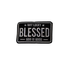 May include: A black and white patch with the words "NOT LUCKY" above "BLESSED" and "GOD IS GOOD" below. The patch has a rectangular shape with a white border and is likely an iron-on or sew-on patch.