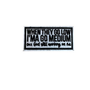 Peut inclure: Un écusson rectangulaire noir et blanc avec le texte "WHEN THEY GO LOW I'MA GO MEDIUM cuz God still working on me." L'écusson a une bordure blanche et est probablement en tissu.