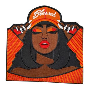 Puede incluir: Parche bordado que presenta un retrato estilizado de una mujer con piel oscura, que lleva una gorra naranja con la palabra "Blessed" y una parte superior a rayas naranja y roja. La mujer tiene los ojos cerrados, labios rojos y pelo largo negro.