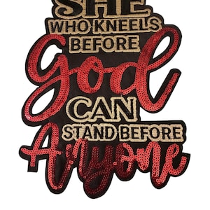 May include: Black patch with gold and red text that reads "SHE WHO KNEELS BEFORE GOD CAN STAND BEFORE ANYONE." The text is embroidered with gold thread and red sequins. The patch is rectangular with rounded edges.